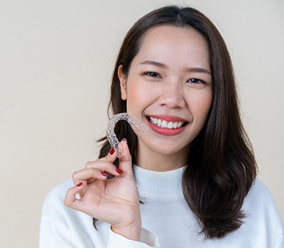 A young woman holding a clear Invisalign aligner in her right hand and smiling