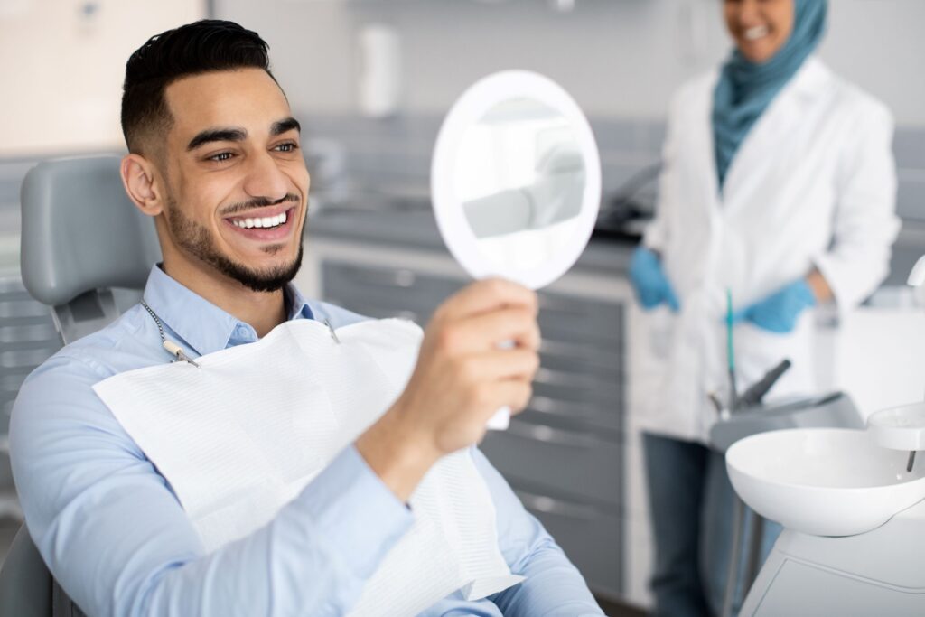 Man in dental chair with dark hair smiling at his reflection in handheld mirror